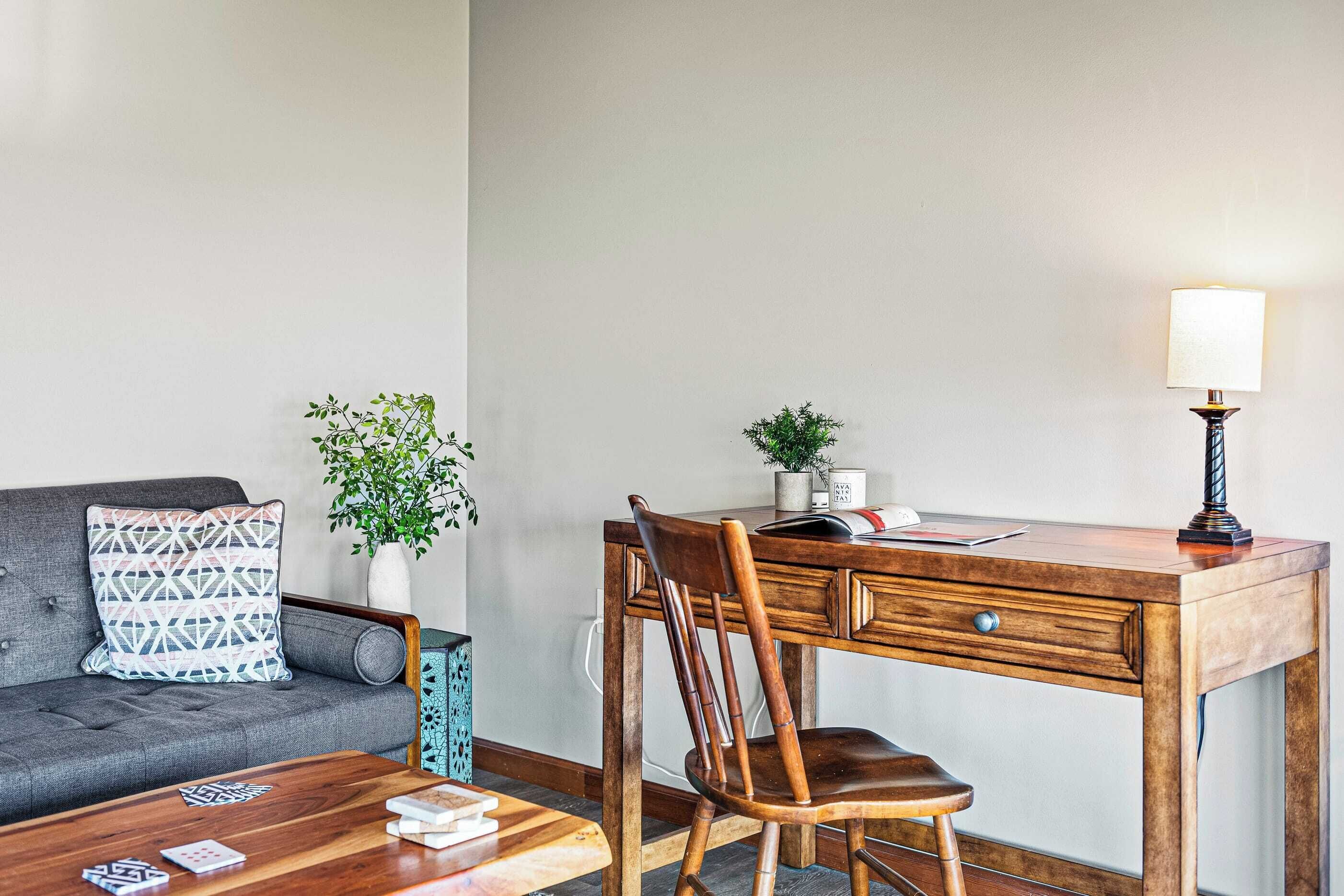 Et værelse med et træskrivebord, en stol, en grå sofa, en pude med mønster, en lille plante og en lampe.