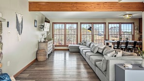 A living room with a sectional sofa, a dining area with chairs, large windows, and a stone wall. - Laker's Pointe by Avantstay Lakefront Private Peninsula Hot Tub, Game Room (Long Pond)