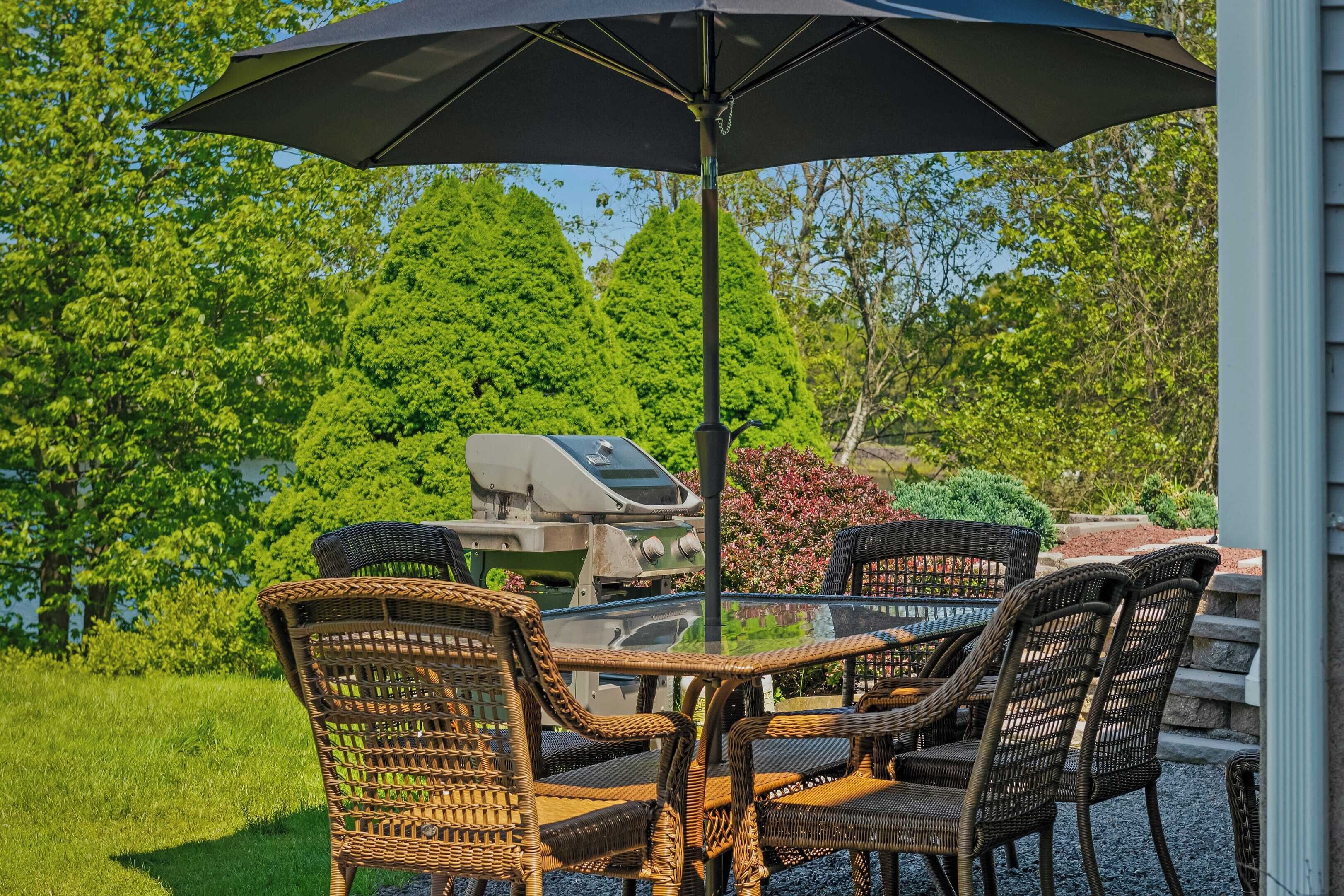 Patio esterno con sedie in vimini e un tavolo sotto un ombrellone grande, affacciato su un giardino rigoglioso e un barbecue.
