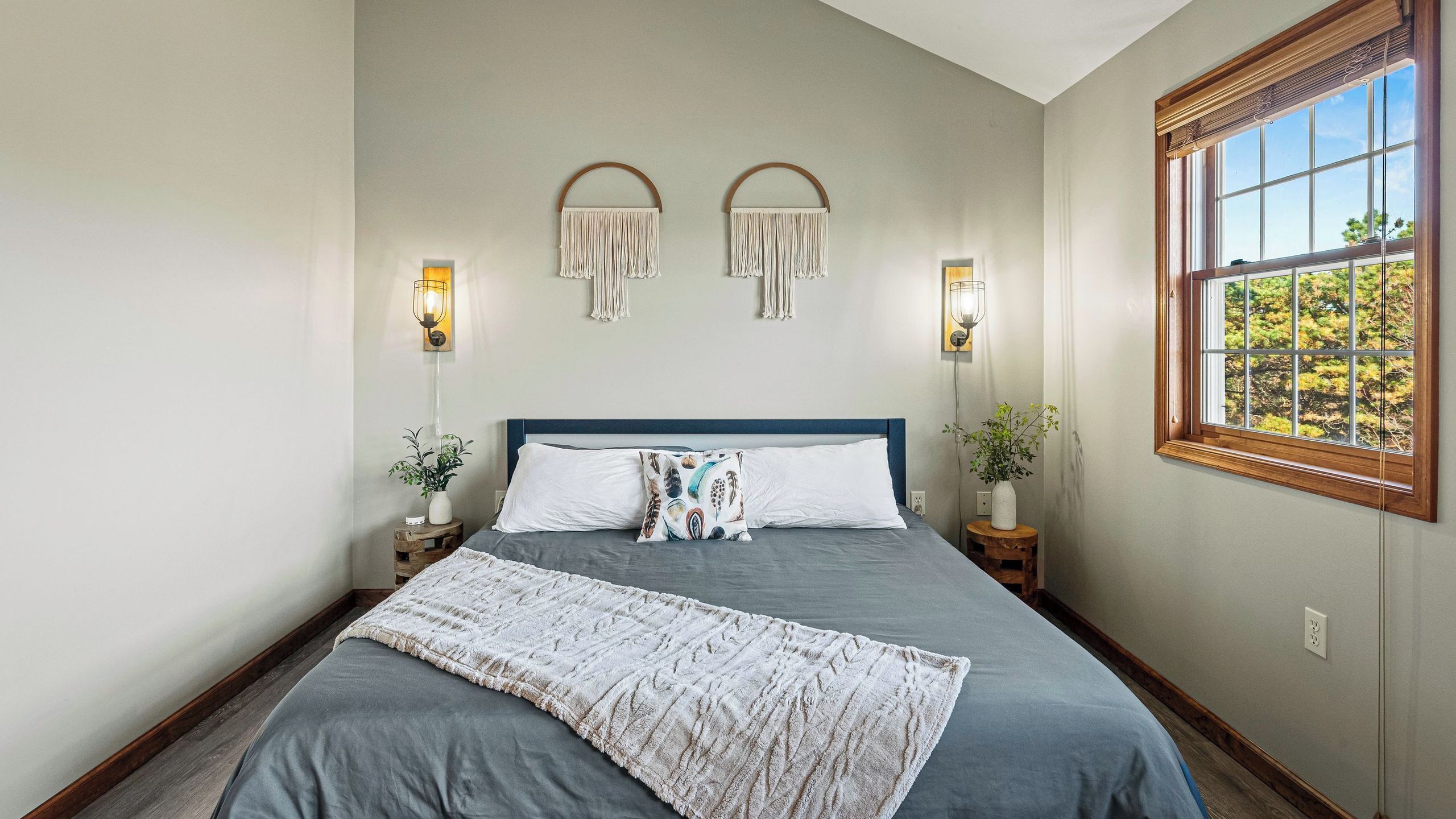 A bedroom with a bed, two wall-mounted lamps, a window with a view, and a decorative wall hanging.