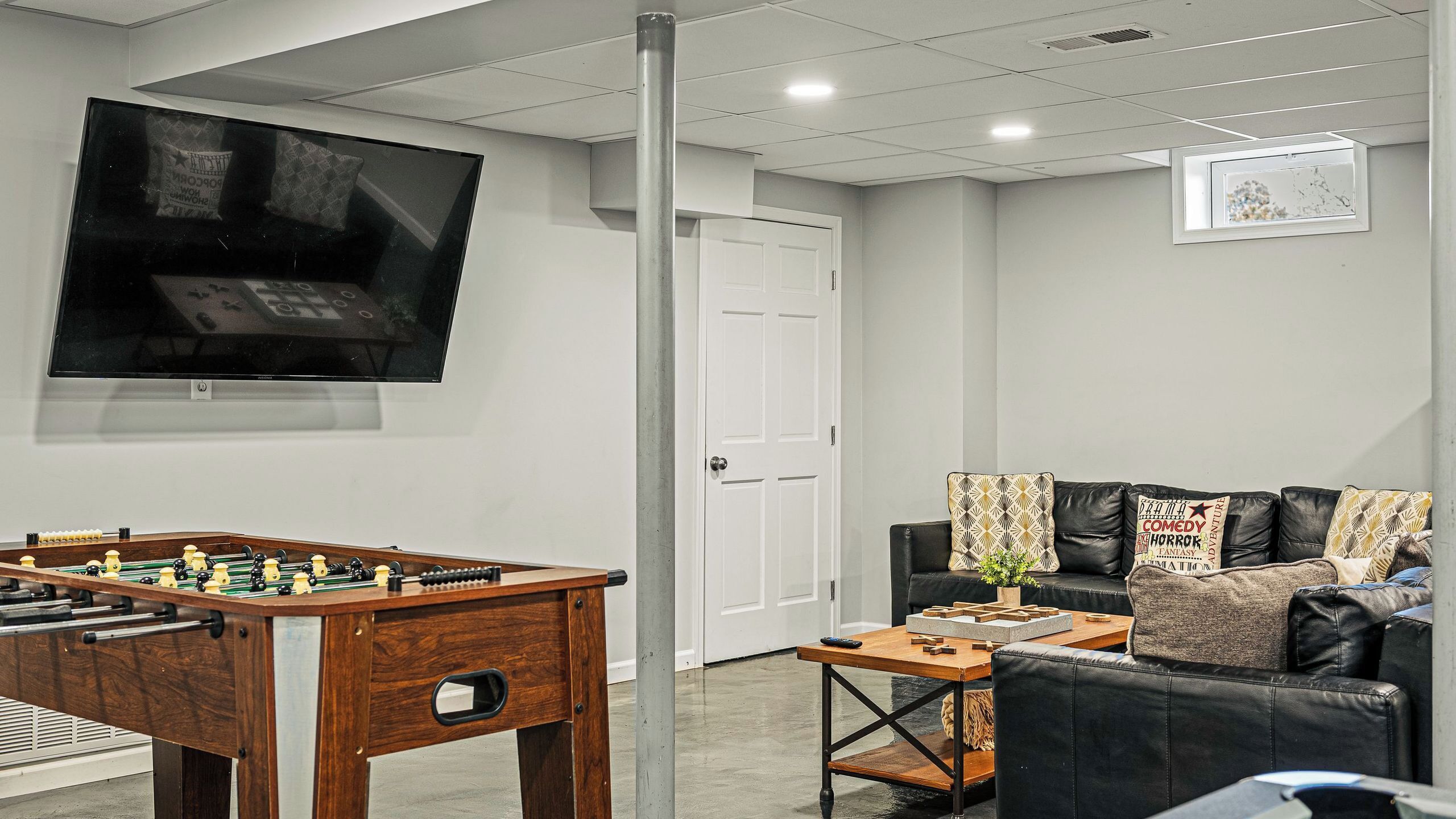 A room with a foosball table, a television mounted on the wall, a couch with patterned cushions, and a door.