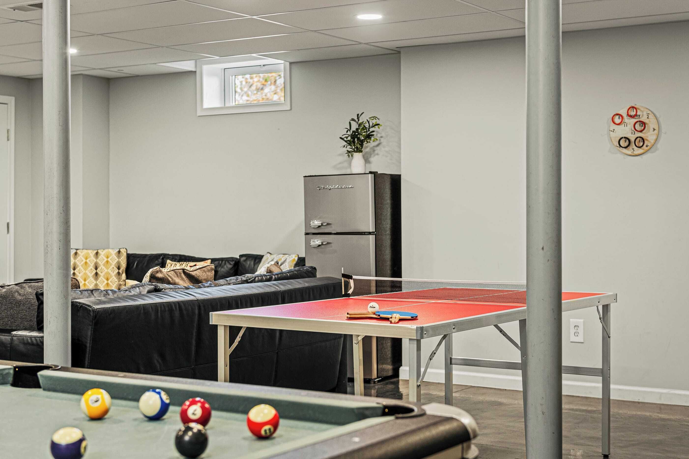 A billiards table, a pool table, and a sofa in a room with a grey wall and a window.