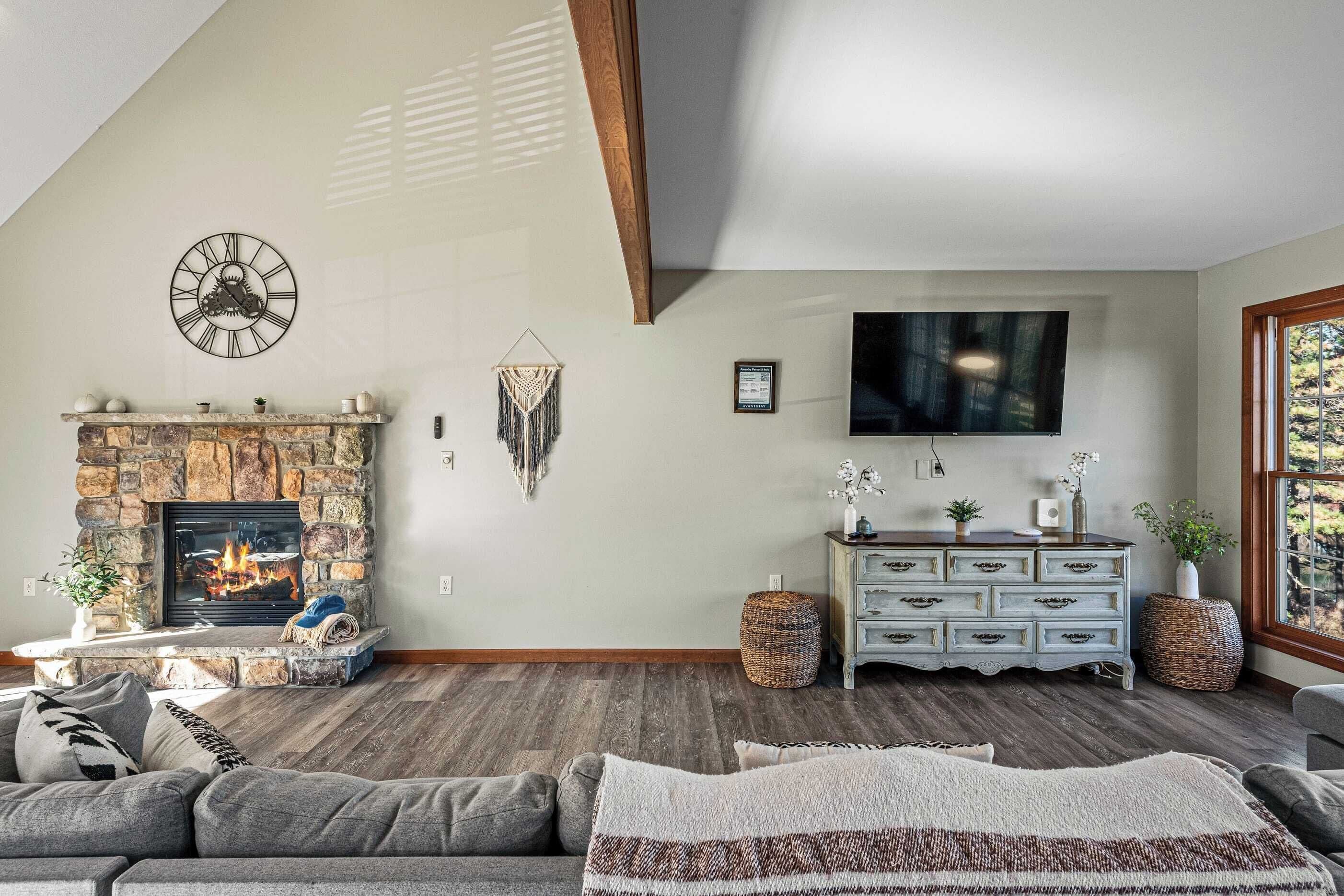 Ein Wohnzimmer mit Kamin aus Stein, einer Uhr, einem Fernseher und einem großen Fenster.