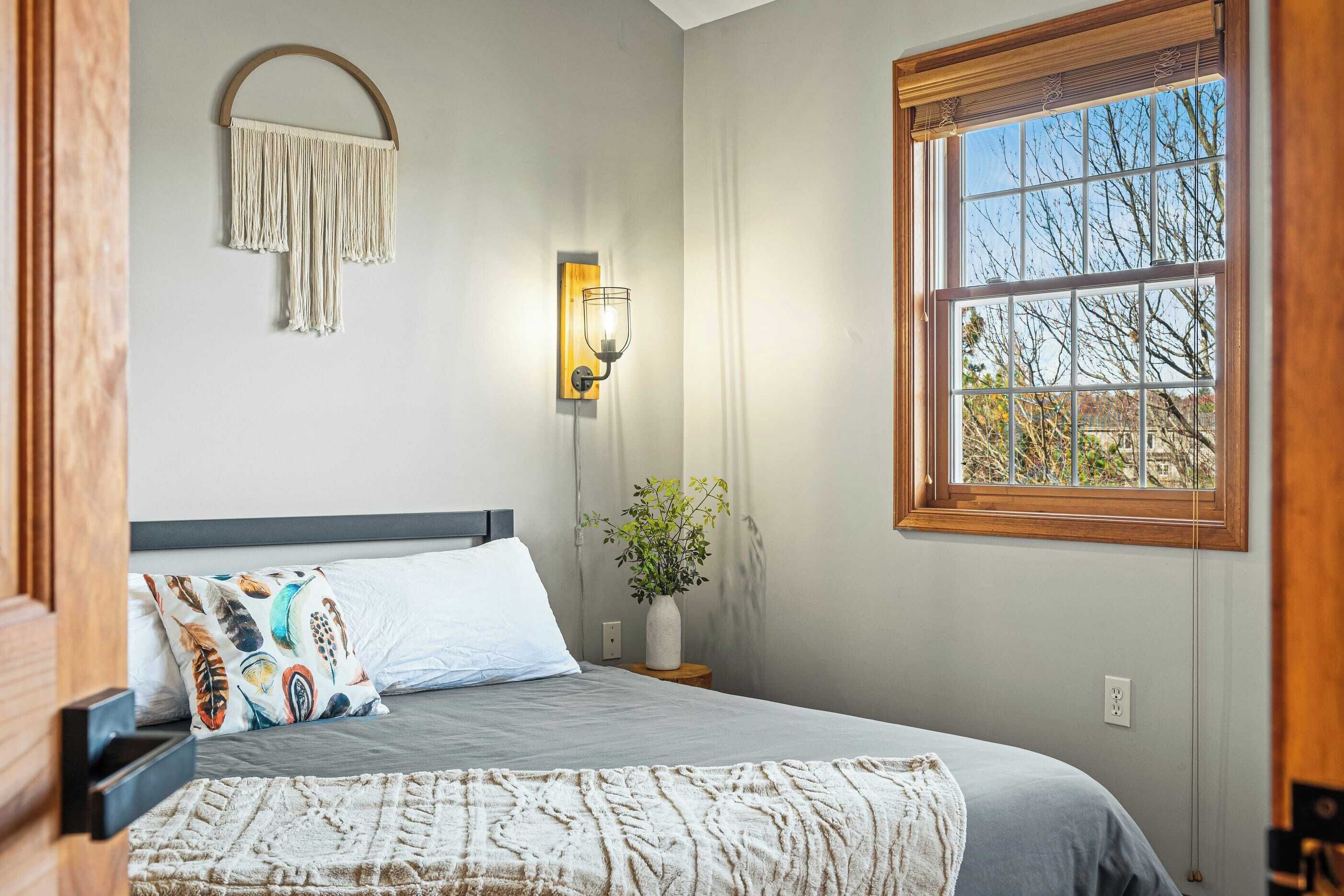 A bedroom with a bed, a wall-mounted macrame light fixture, a window with a view of trees, and a bedside plant.