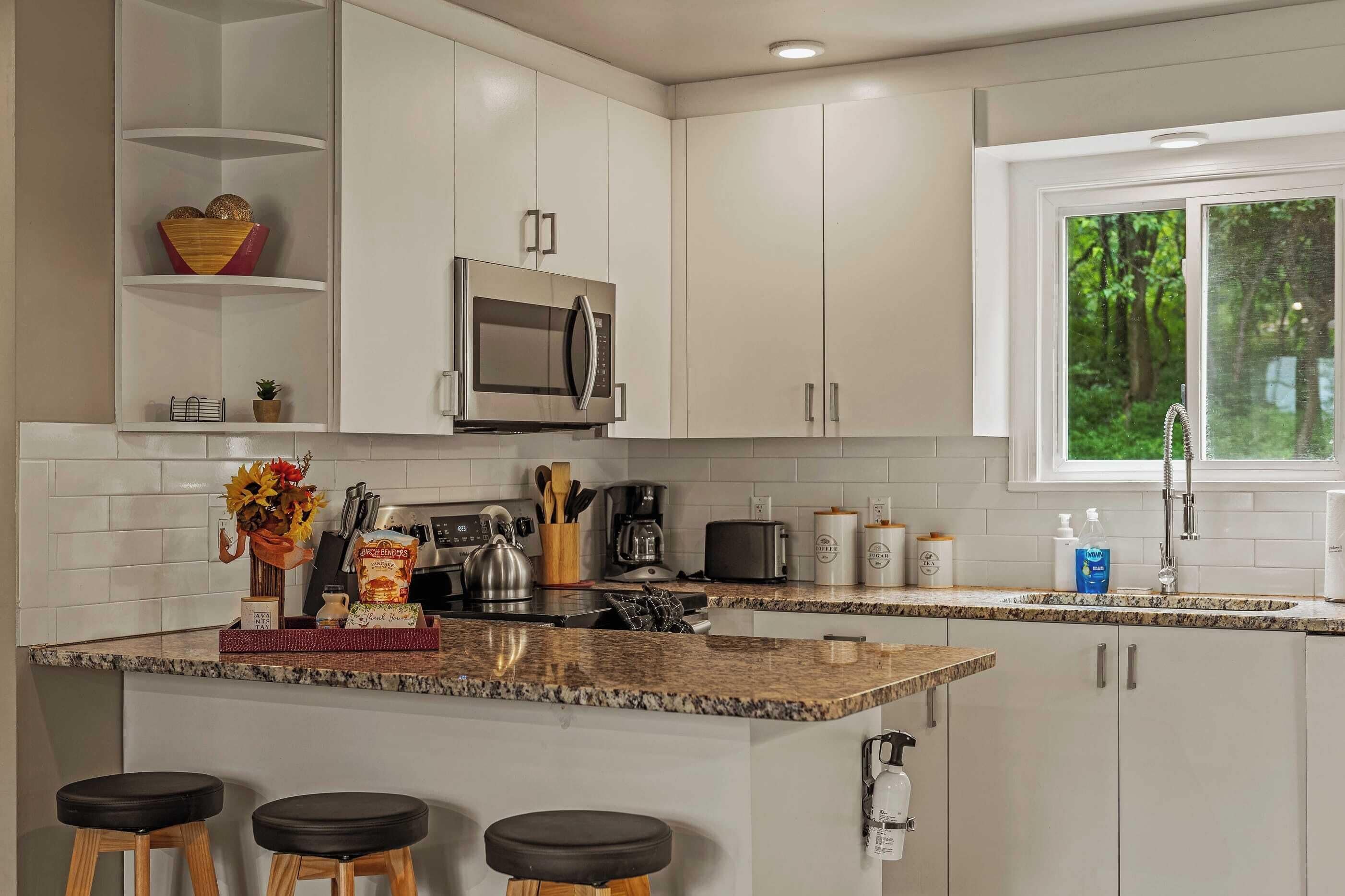 Una cocina moderna con armarios blancos, un microondas de acero inoxidable, una cafetera y un ventanal con vistas a árboles.