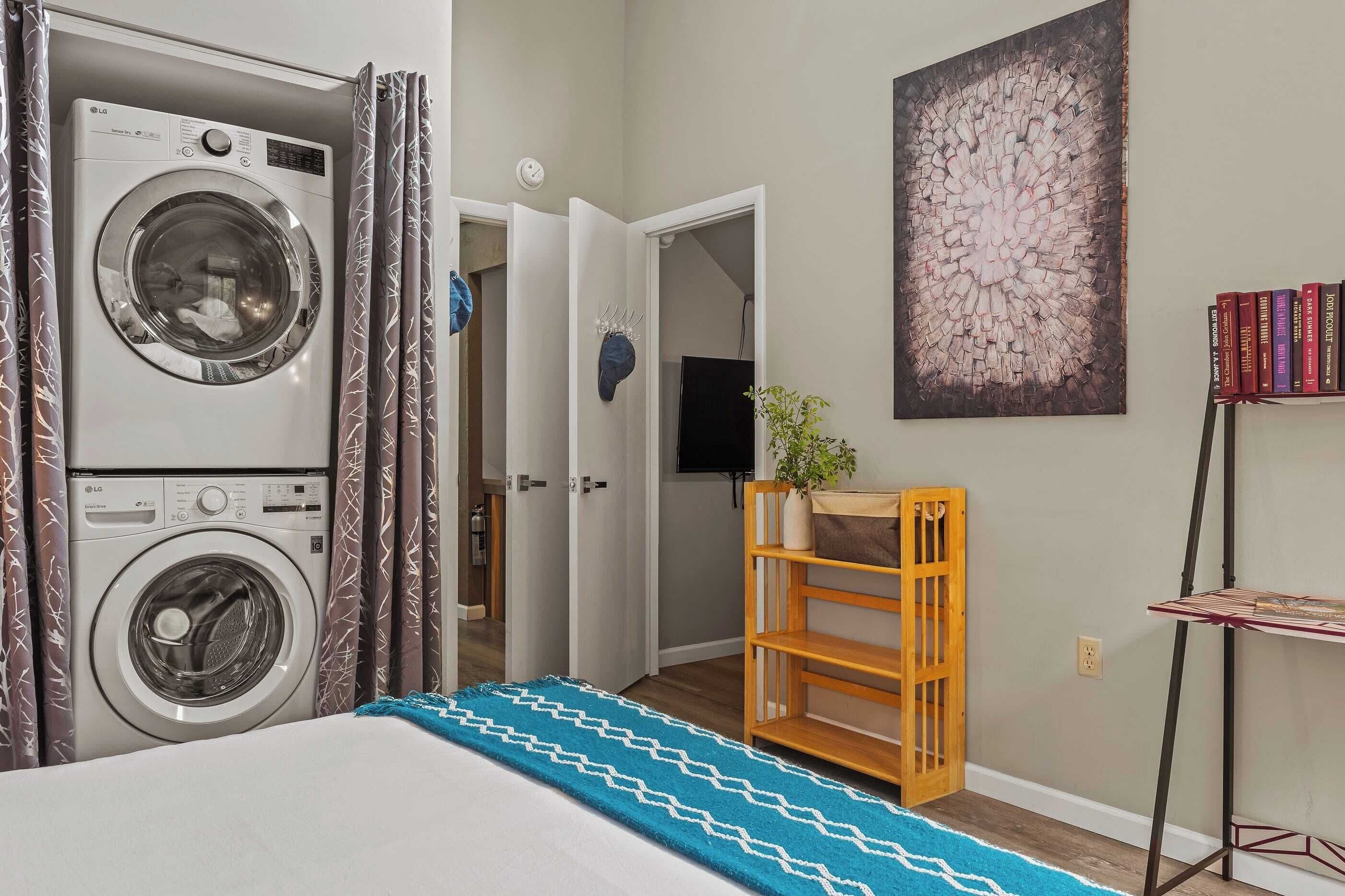 A laundry room with a front-loading washing machine, a wooden shelf with books, and a painting on the wall.