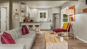 Un salon moderne avec un canapé gris, des coussins rouges, une table basse en bois et un fauteuil rouge. On aperçoit, à l’arrière-plan, une cuisine avec des meubles blancs et une fenêtre.