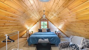 A cozy attic bedroom with a sloped ceiling, a bed with a blue cover, a chair, a nightstand, and a window with a view of trees.