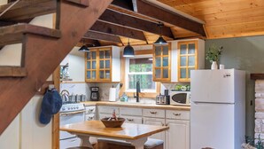 A rustic kitchen with wooden beams, a wooden table with stools, white cabinets, and a refrigerator.