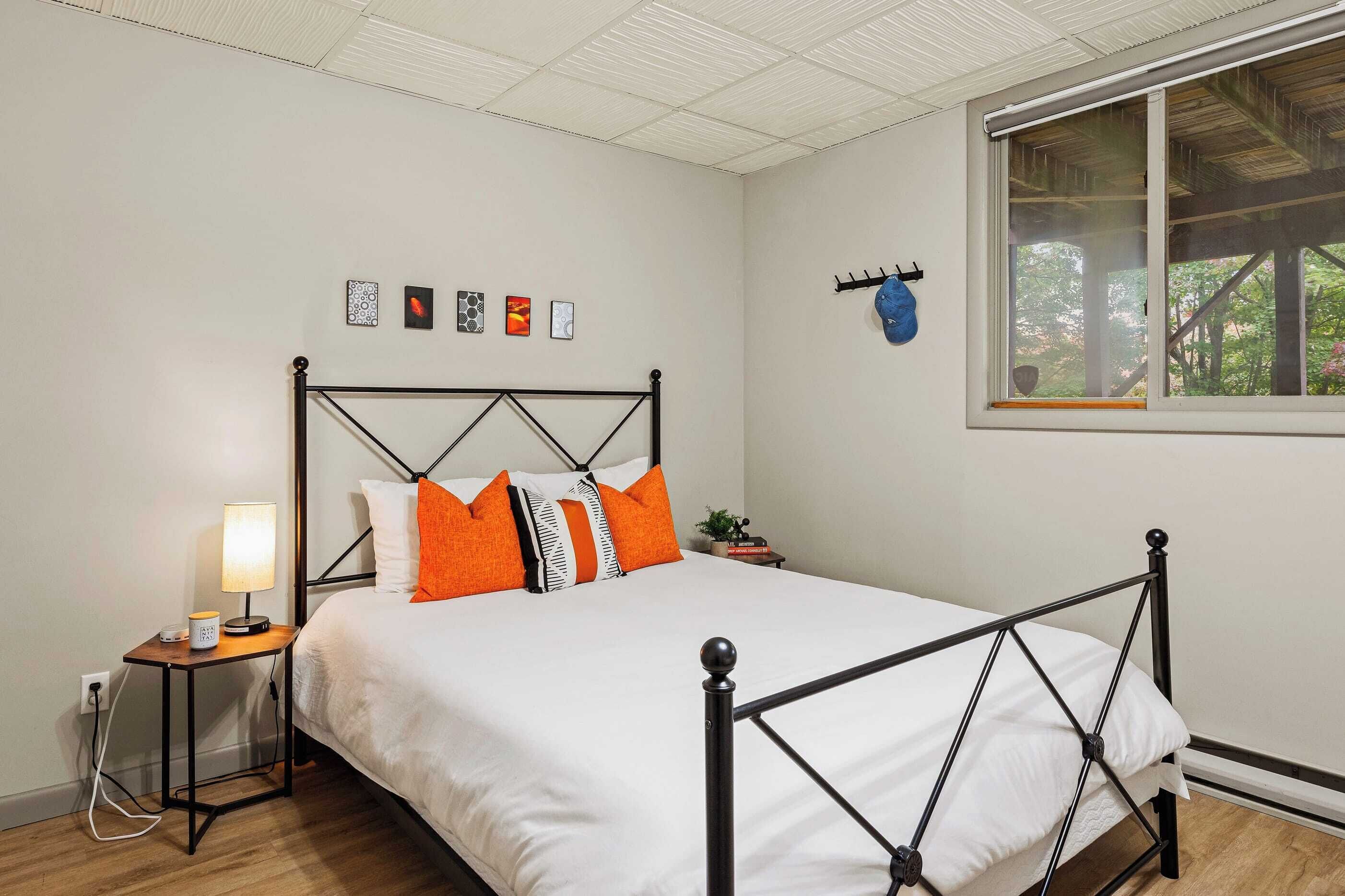 Un lit bien fait, avec un cadre en métal, des draps blancs et des oreillers orange. On y trouve une table de chevet avec une lampe et une petite plante. La pièce a des murs blancs, une fenêtre donnant sur des arbres et un plafond orné d’un motif en réseau.