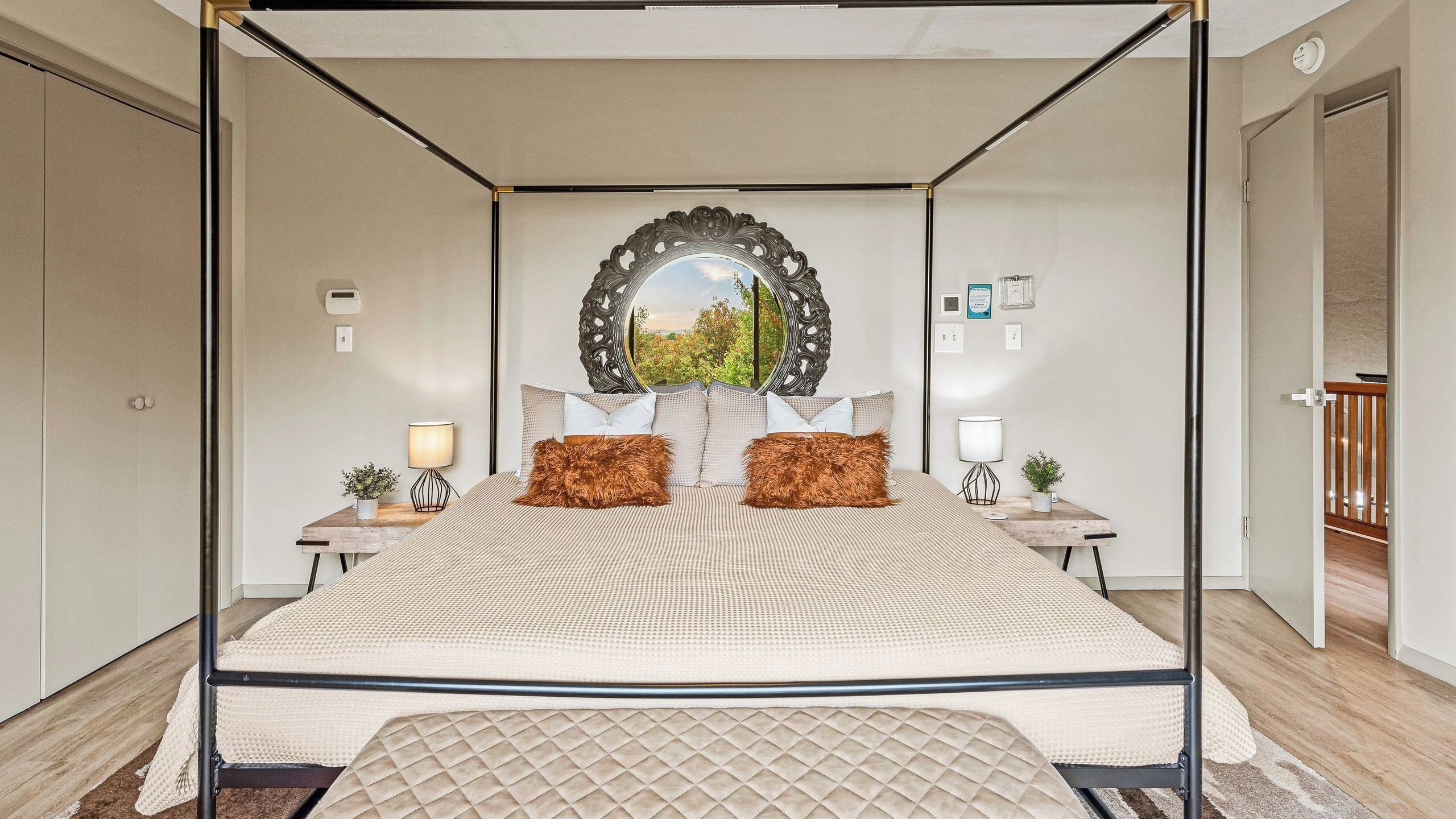 A four-poster bed with a quilted headboard, two nightstands with lamps, a mirror above the bed, and a view of greenery outside.