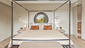 Un lit à baldaquin avec une tête de lit matelassée, deux tables de chevet avec des lampes, un miroir au-dessus du lit et une vue sur la verdure à l’extérieur.
