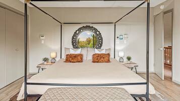 A four-poster bed with a quilted headboard, two nightstands with lamps, a mirror above the bed, and a view of greenery outside.
