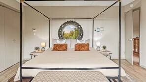 Uma cama com dossel e cabeceira acolchoada, duas mesas de cabeceira com abajures, um espelho sobre a cama e vista para o verde lá fora.