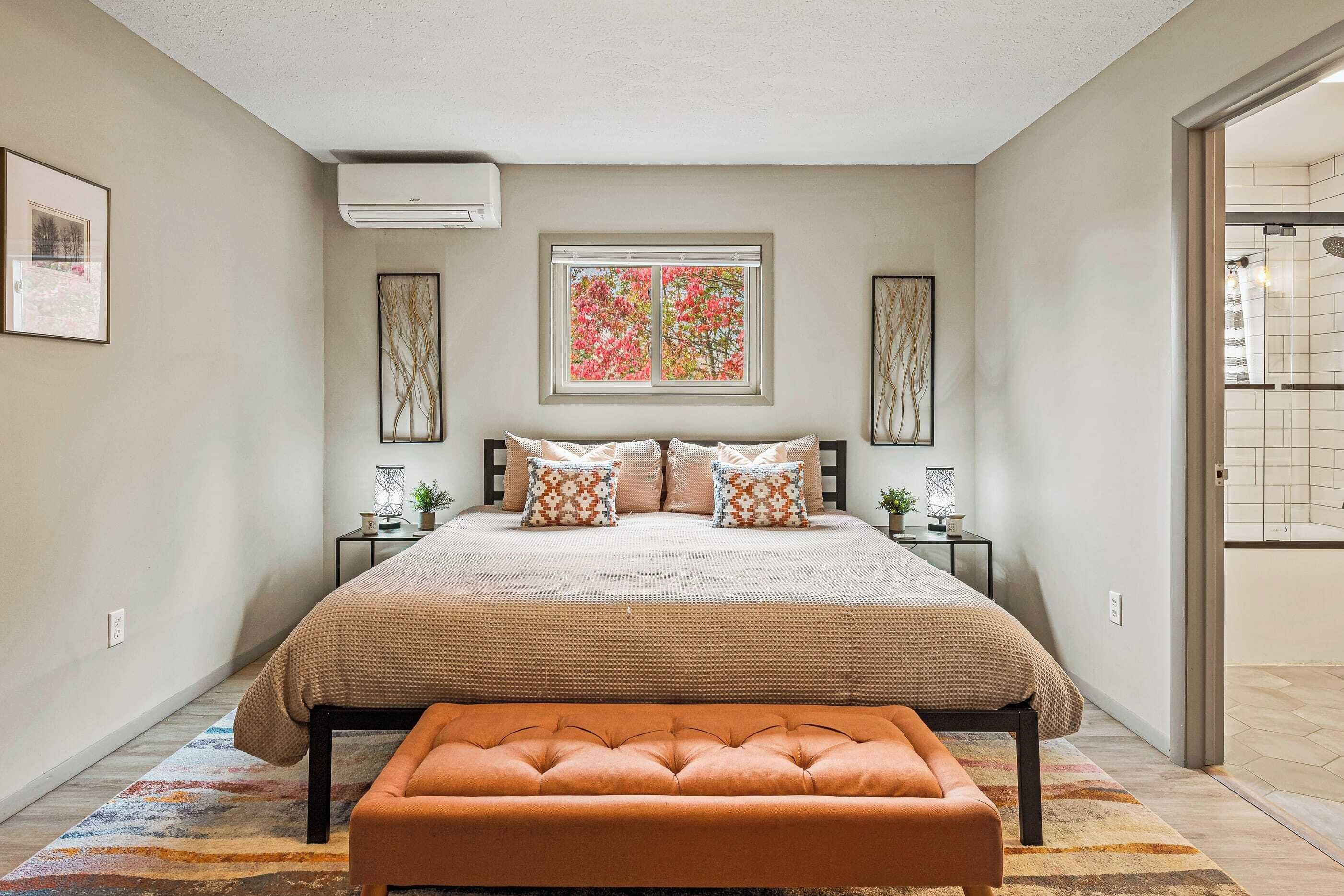 A bedroom with a large bed, a bench, a window with a view, and a door leading to a bathroom.