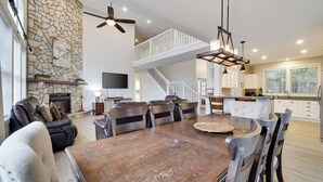 A spacious living area with a stone fireplace, a dining table, and a kitchen with white cabinets. - The Nest at Pinecrest by Avantstay Spacious Deck, Game Room, Fire Pit (Pocono Pines)