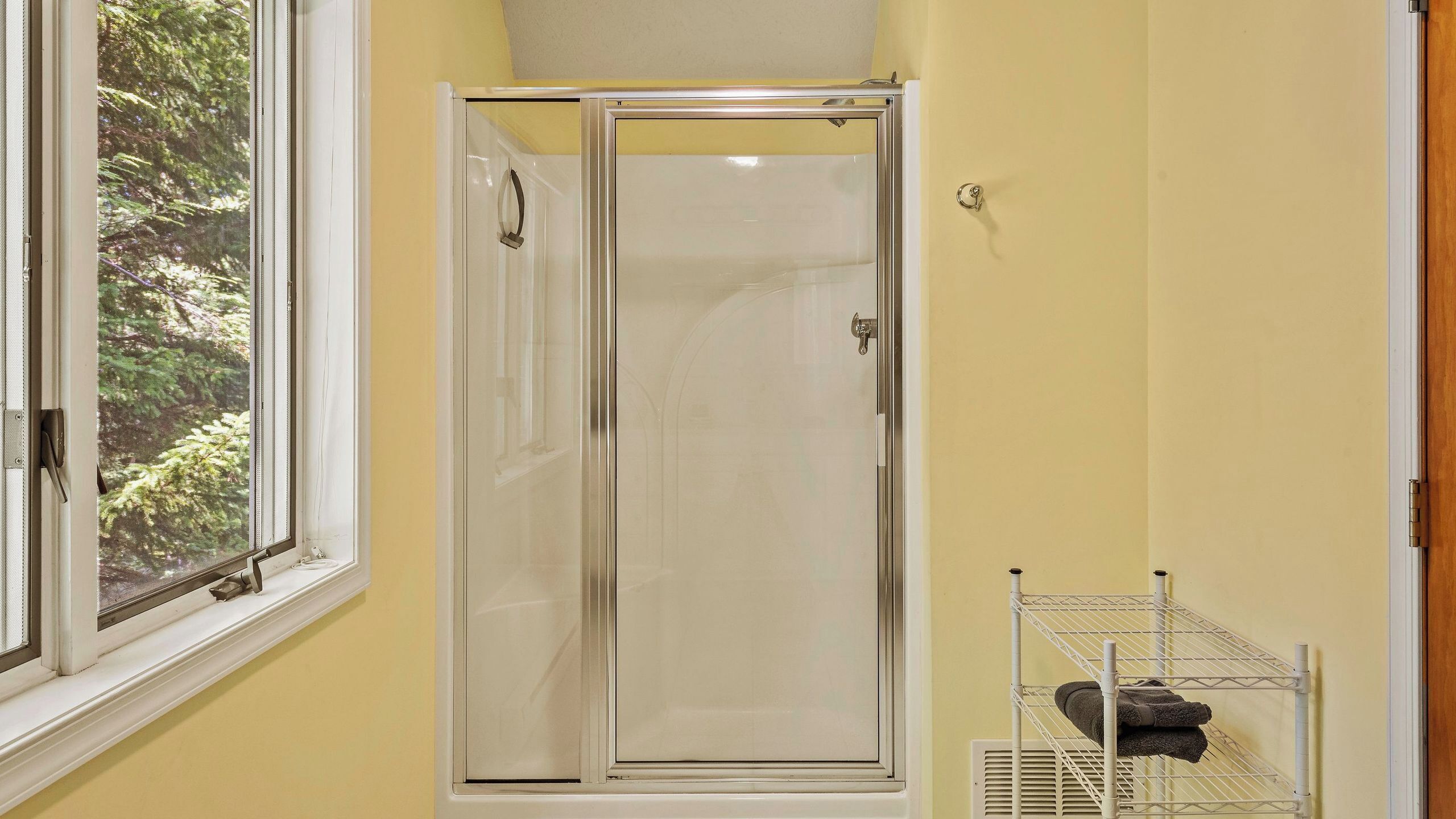 A bathroom with a glass shower enclosure, a window with a view of trees, and a towel rack with towels.