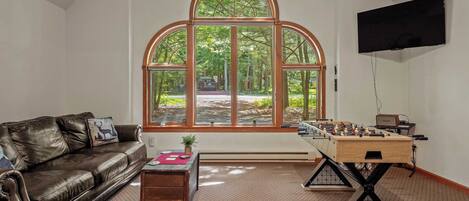 A living room with a large arched window, a leather sofa, a foosball table, and a flat-screen TV.