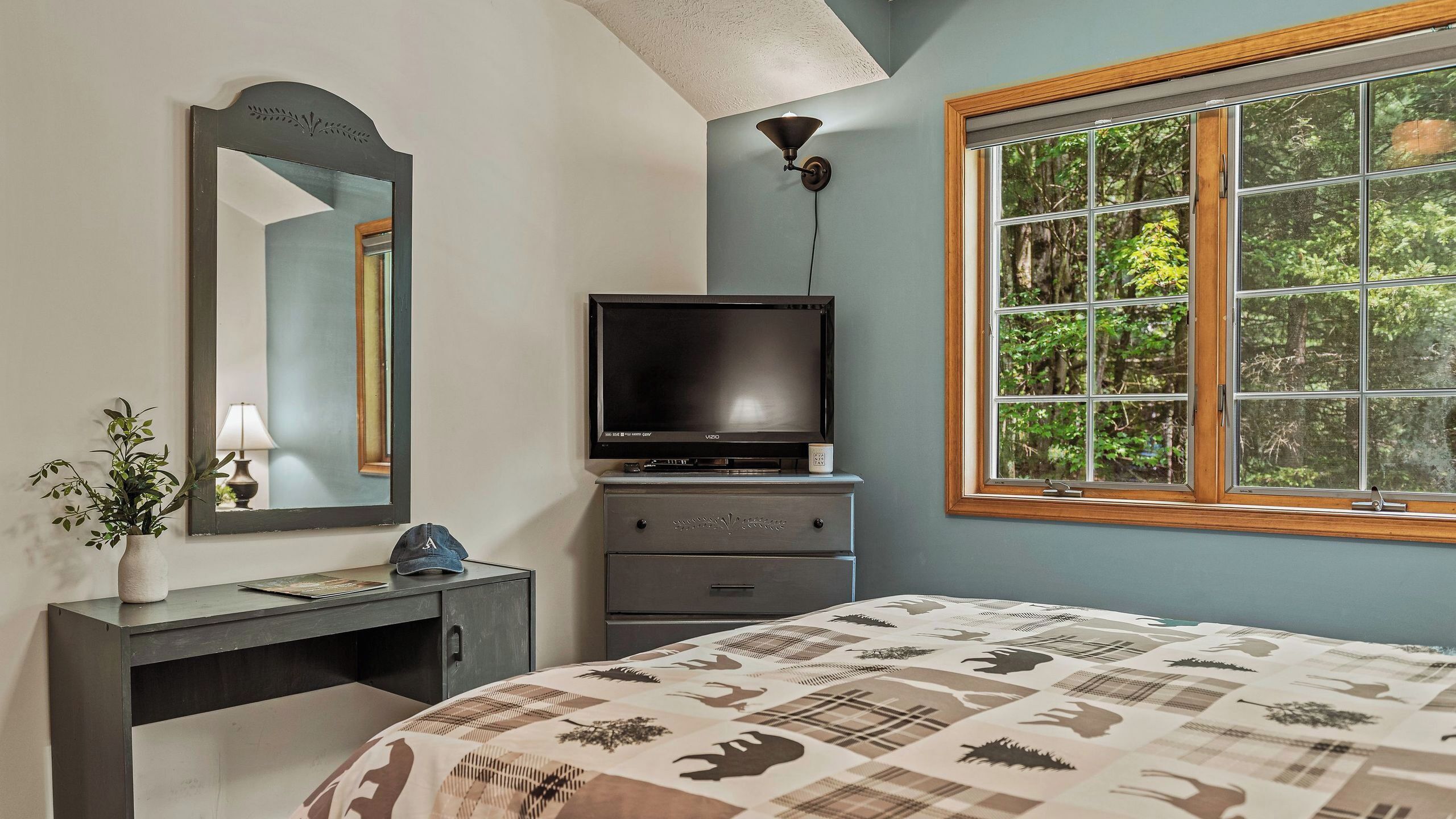 Un dormitorio con cama, tocador, televisor, espejo y ventana con vistas a árboles.