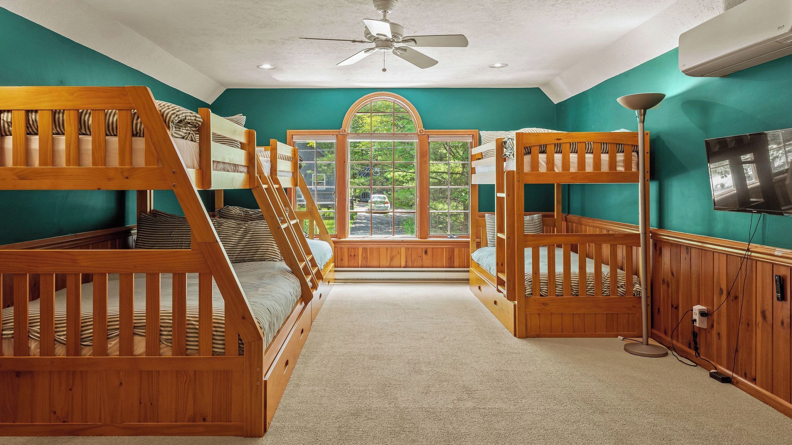 Une chambre avec des lits superposés en bois, un ventilateur de plafond, une grande fenêtre en arc de cercle et un téléviseur.