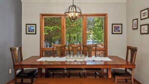 Ein Esszimmer mit einem großen Holztisch, sechs Stühlen, einer Kandelaber und großen Fenstern mit Blick auf Bäume und ein Gewässer.