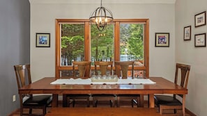 A dining room with a large wooden table, six chairs, a chandelier, and large windows with a view of trees and a body of water.