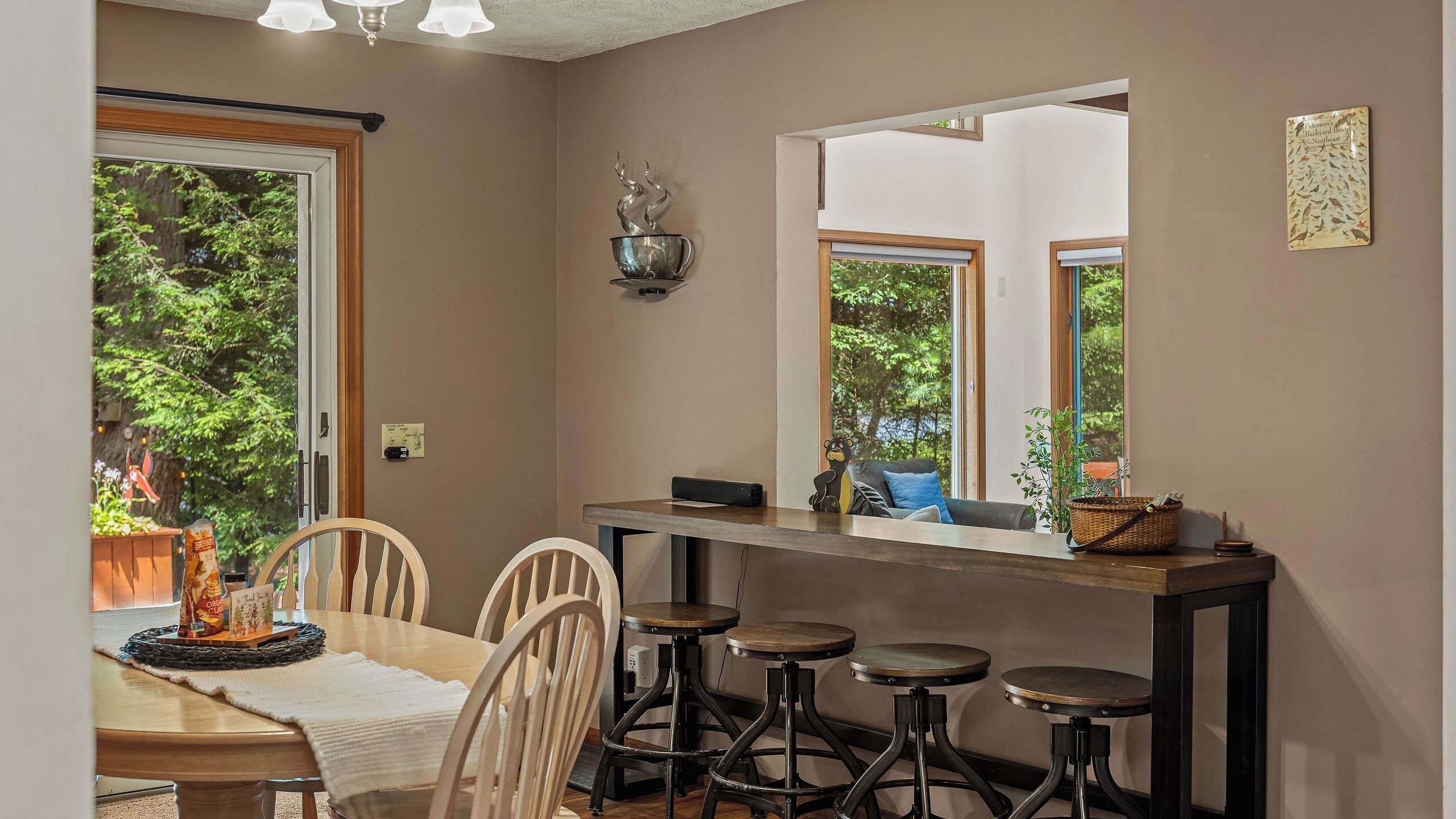 A dining area with a wooden table, four stools, a large mirror, and a view of greenery outside.