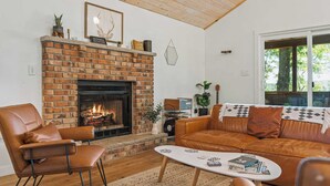 A cozy living room with a brick fireplace, leather furniture, and a view of trees through a large window. - Lake Daze by Avantstay Lakefront w/ Modern Interior, Enclosed Porch & Hot Tub (Pocono Summit)