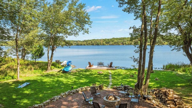 A lakeside deck with outdoor seating, a fire pit, and a view of the lake. - Lake Daze by Avantstay Lakefront w/ Modern Interior, Enclosed Porch & Hot Tub (Pocono Summit)