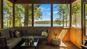 A cozy room with a wooden floor, a sofa with cushions, a coffee table with a plant, and large windows offering a view of trees and a lake. - Lake Daze by Avantstay Lakefront w/ Modern Interior, Enclosed Porch & Hot Tub (Pocono Summit)