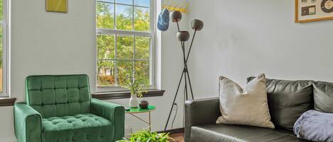 A living room with a green armchair, a leather sofa, a coffee table, and a window with a view of trees.