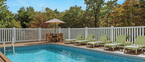 A swimming pool with a wooden deck, white picket fence, and green lounge chairs.