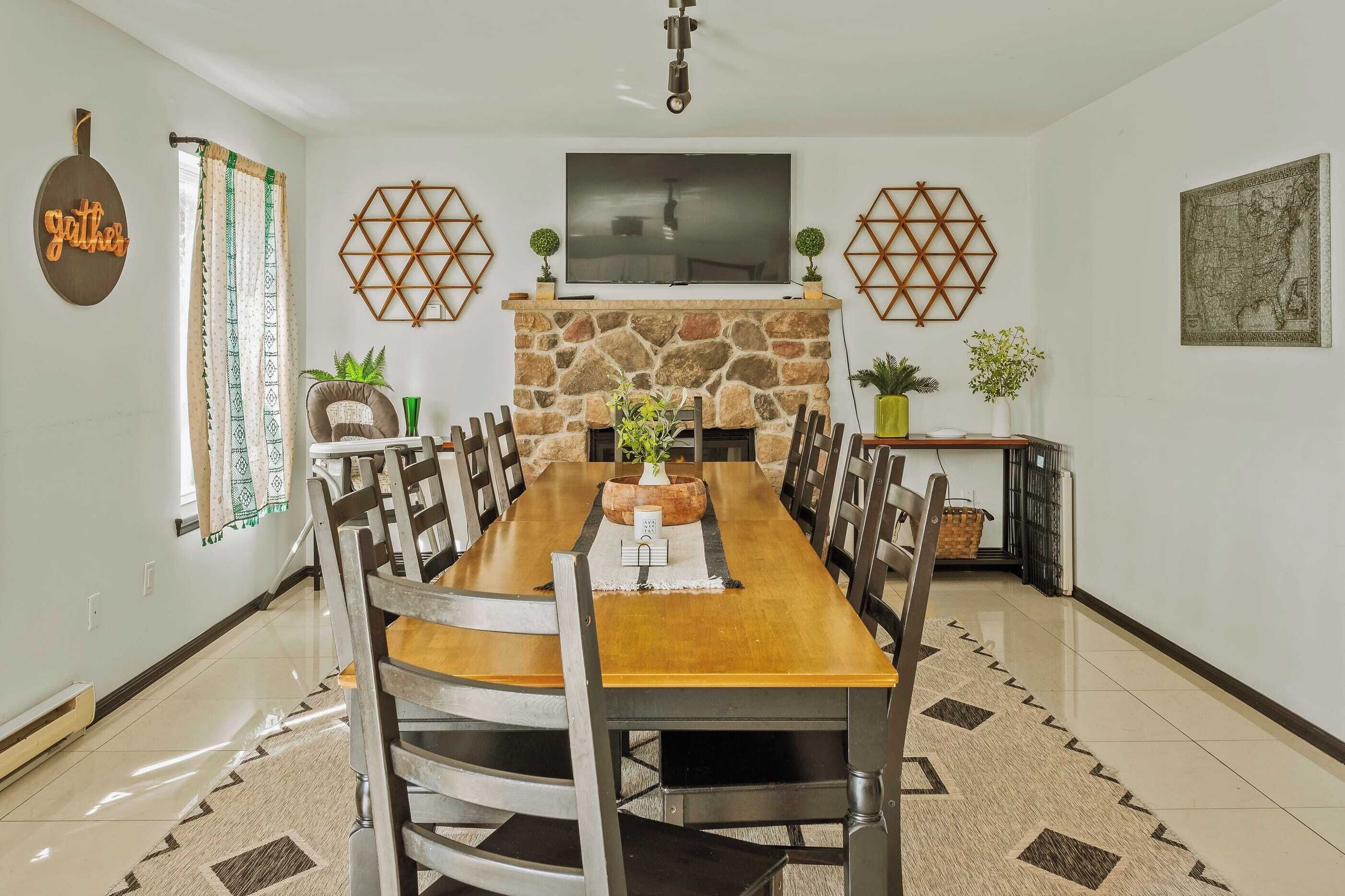 A dining area with a long wooden table, chairs, a stone fireplace, a television, decorative wall art, and a 'Gather' sign.