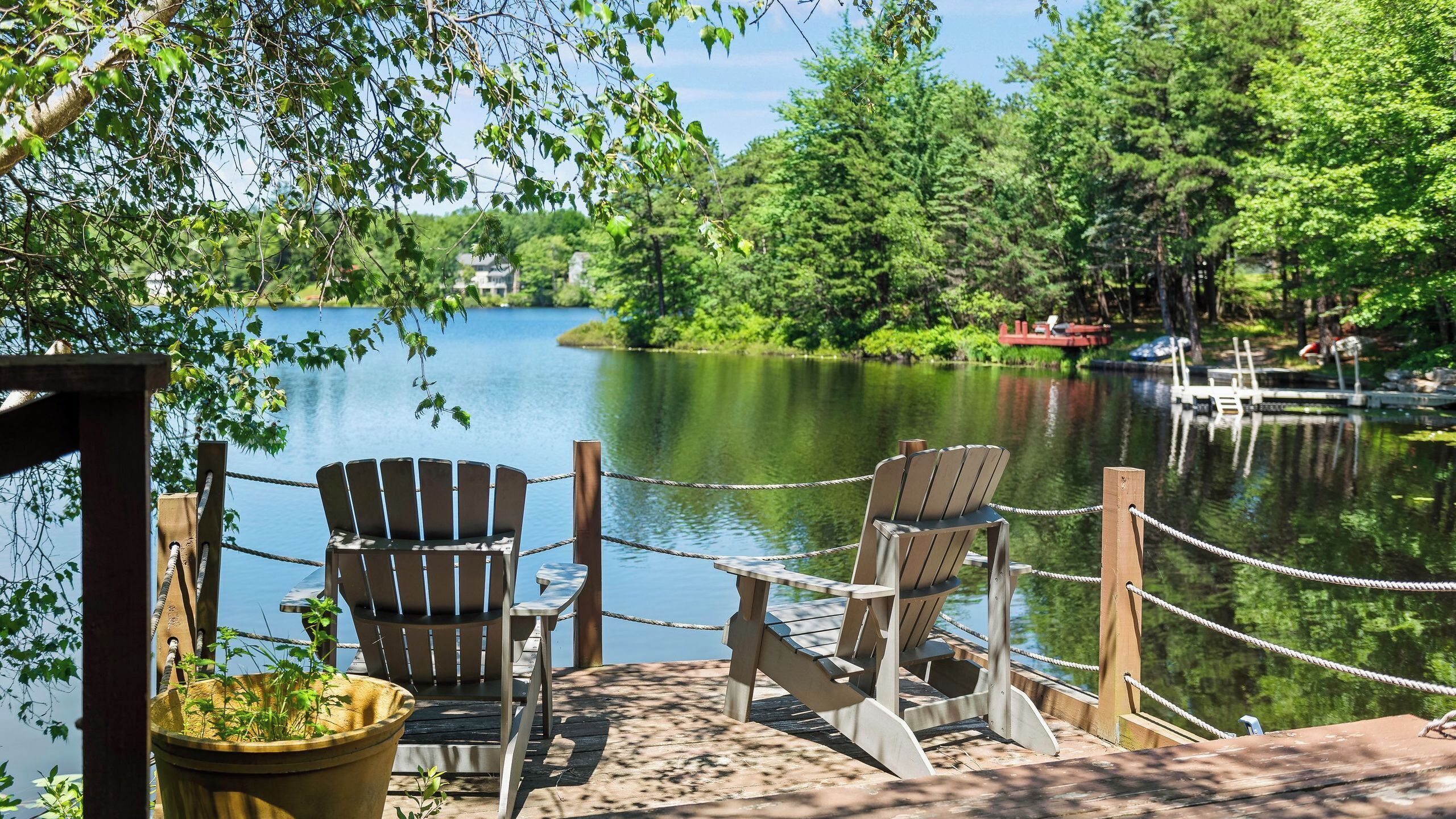 A wooden dock with two Adirondack chairs facing a calm lake, surrounded by lush greenery and a distant red bridge.