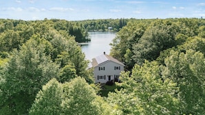 Una casa blanca con techo inclinado, rodeada de árboles verdes frondosos y un lago al fondo.