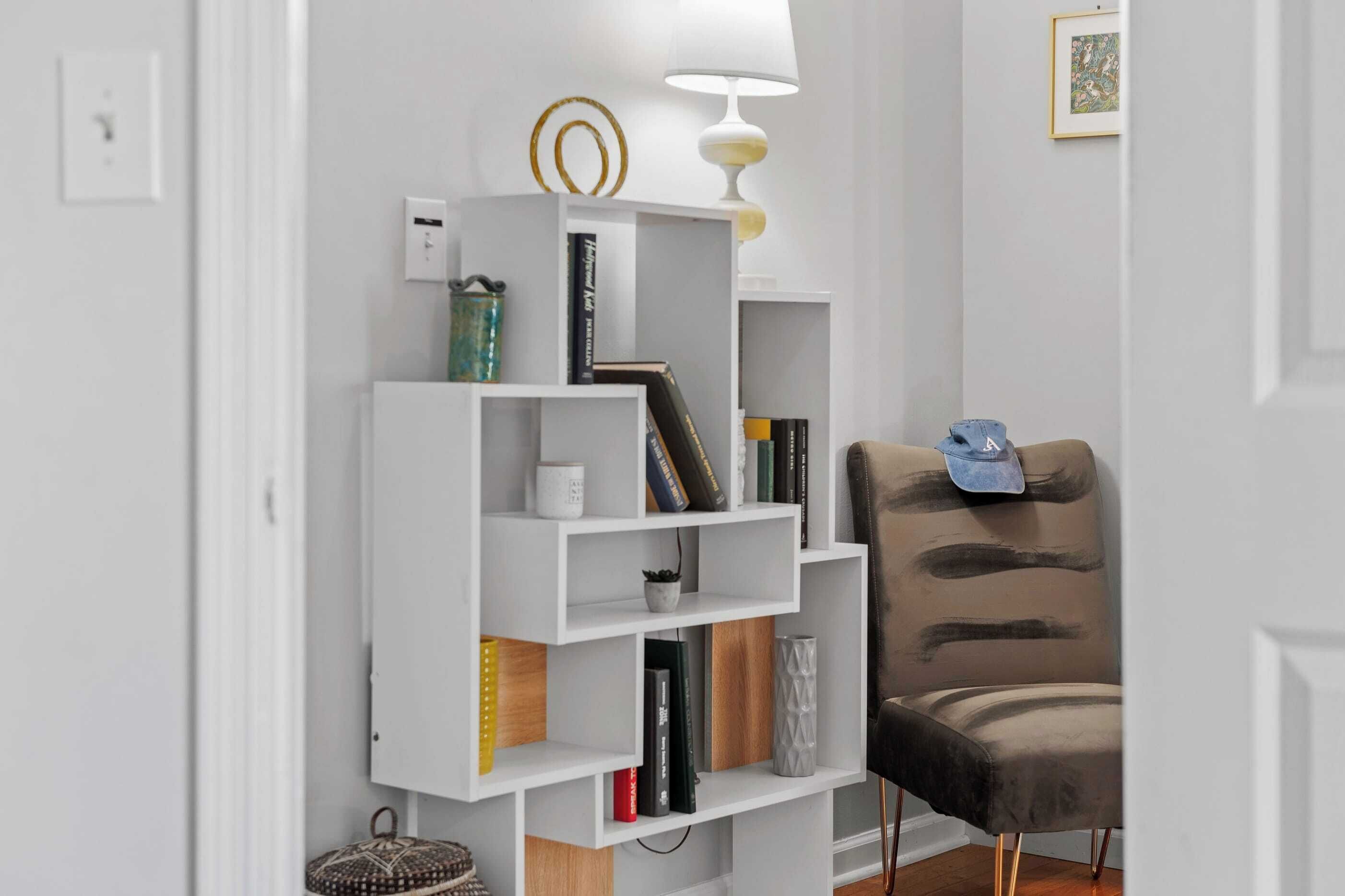 A white bookshelf with books and decorative items, a lamp, and a chair with a unique design.