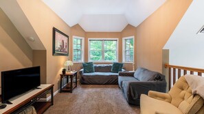 A cozy living room with a TV, sofa, and armchair, a fireplace, and a large window with a view of trees. - Bear Trail Chalet by Avantstay Retreat w/ Outdoor Living Deck & Fire Pit (Pocono Lake)