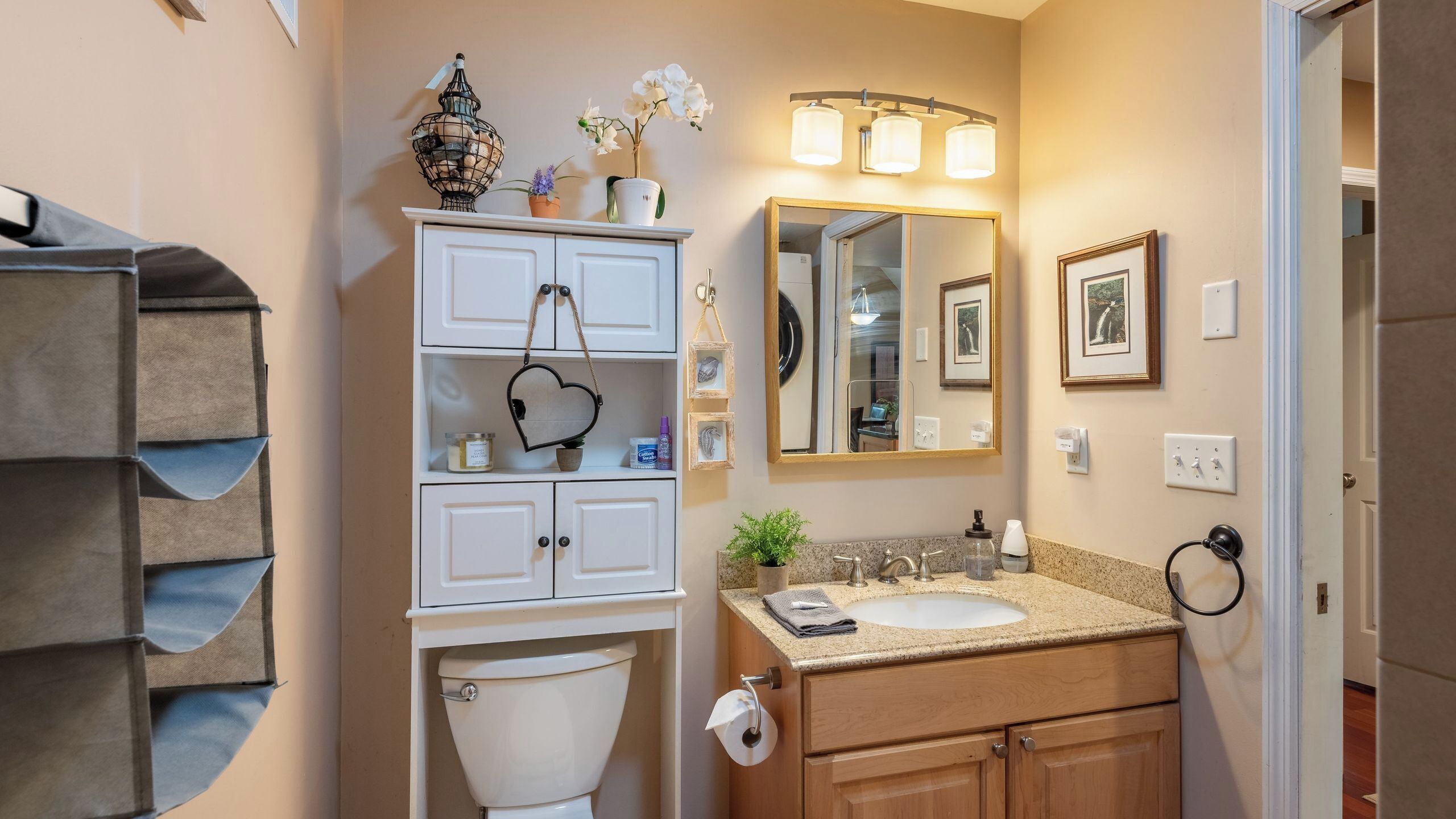 A bathroom with a toilet, sink, vanity, mirror, and shelves with toiletries.