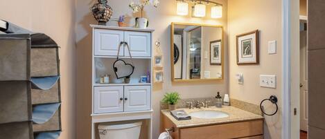 A bathroom with a toilet, sink, vanity, mirror, and shelves with toiletries.