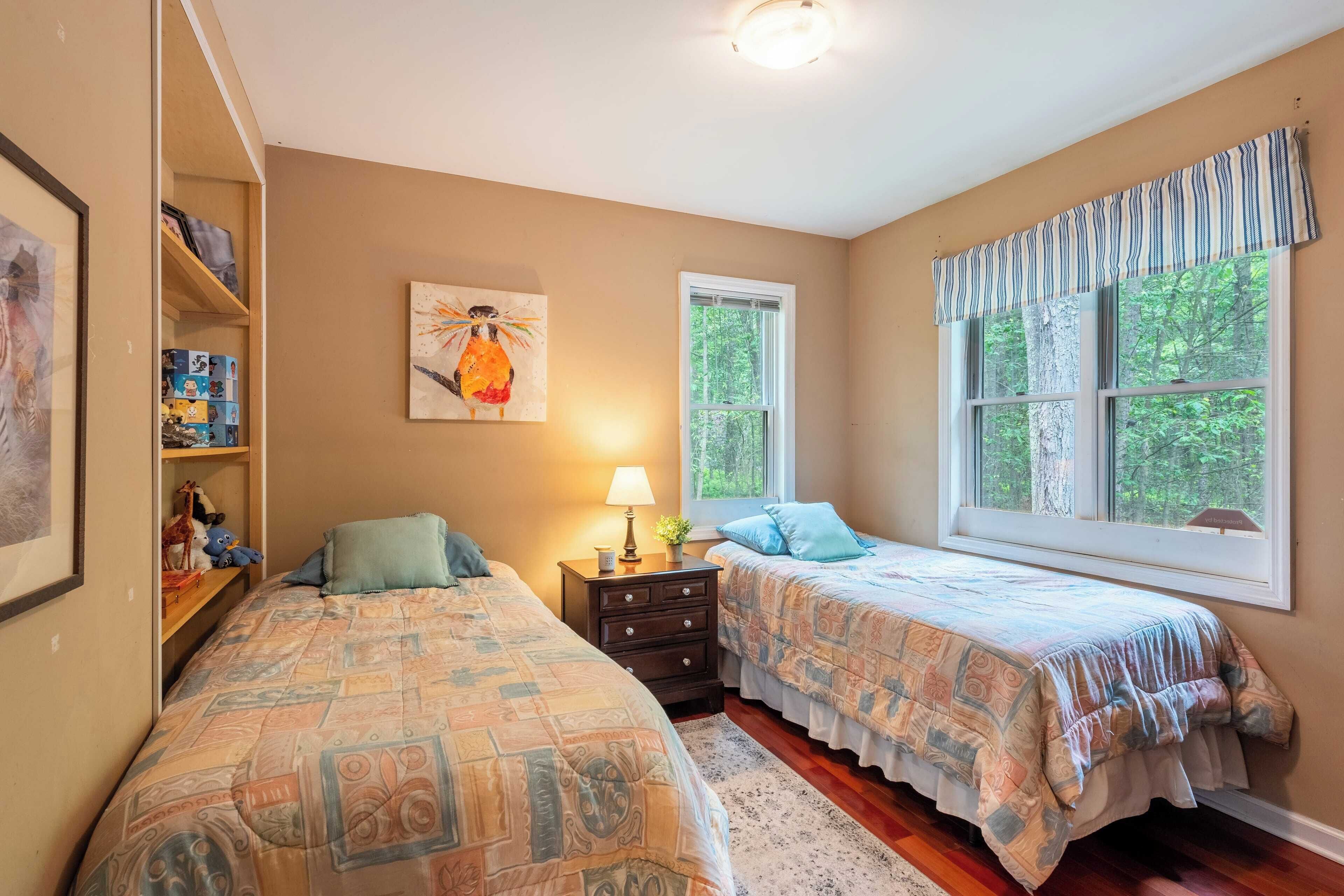 A bedroom with two beds, a nightstand with a lamp, a shelf with stuffed animals, and a window with a view of trees.