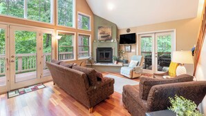 A spacious living room with a fireplace, large windows, and a ceiling fan.