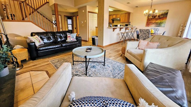 A spacious living room with a black leather sofa, beige armchairs, and a round coffee table. A staircase leads to an upper level, and a kitchen is visible in the background. - Summit Serenity by Avantstay Central Location w/ Mountain Views & Deck (Tannersville)