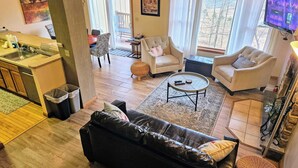 A living room with a black leather sofa, two white armchairs, a coffee table, and a TV mounted on the wall. - Summit Serenity by Avantstay Central Location w/ Mountain Views & Deck (Tannersville)
