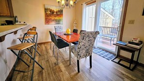 A dining area with a wooden table, chairs, and a chandelier. There is a painting on the wall and a view of the outside through a glass door. - Summit Serenity by Avantstay Central Location w/ Mountain Views & Deck (Tannersville)