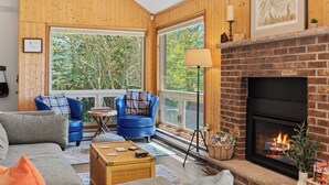 A cozy living room with a fireplace, blue armchairs, and a large window offering a view of trees. - Waterside Haven by Avantstay Lakefront, Hot Tub, Game Room, Fire Pit (Long Pond)