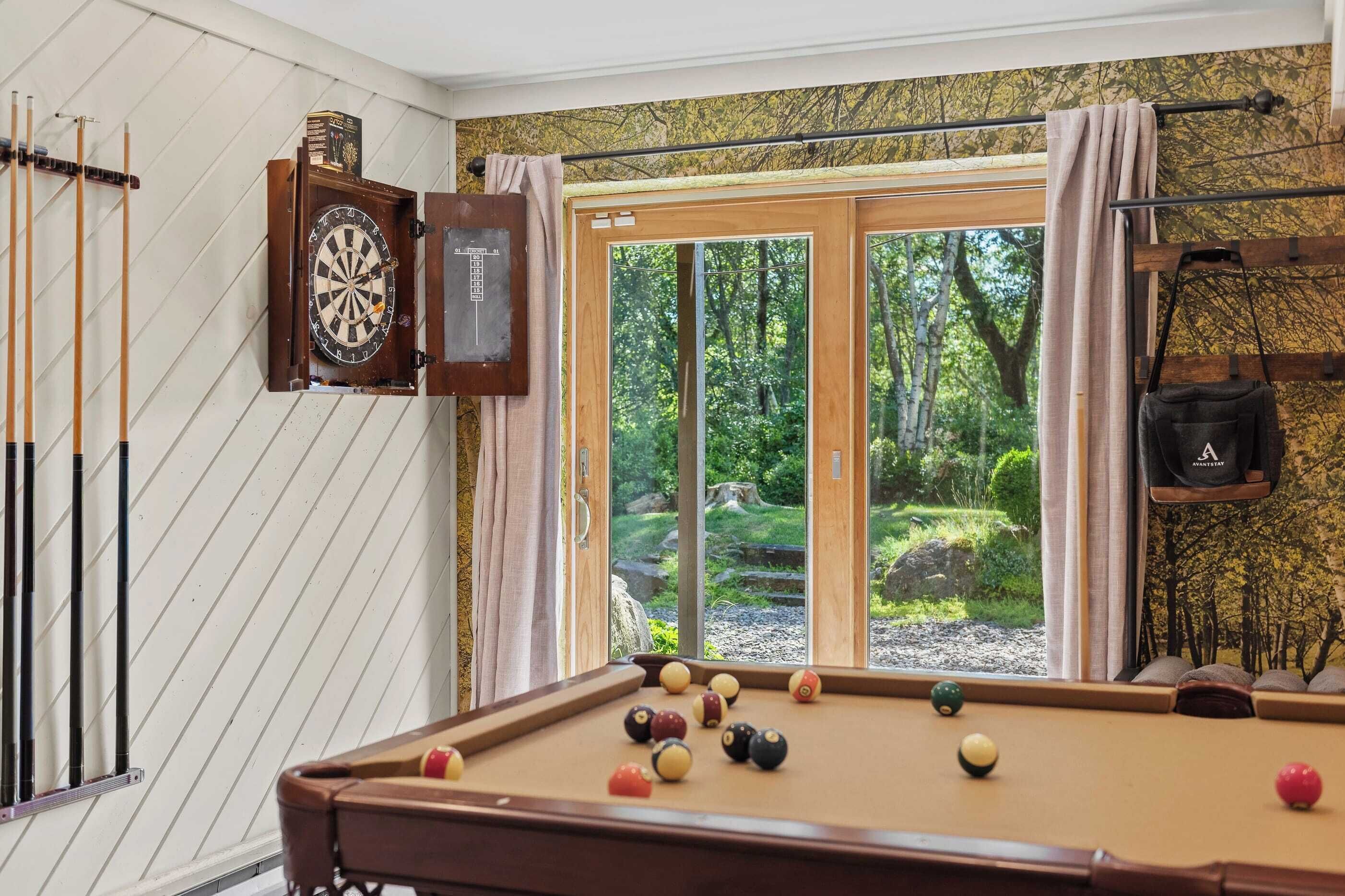 Ein Billardtisch mit Kugeln, eine Dartscheibe und Billardstöcke in einem Raum mit großem Fenster mit Blick auf einen Wald.