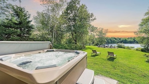 A hot tub with a grassy area and chairs, overlooking a lake with trees and a sunset. - Waterside Haven by Avantstay Lakefront, Hot Tub, Game Room, Fire Pit (Long Pond)