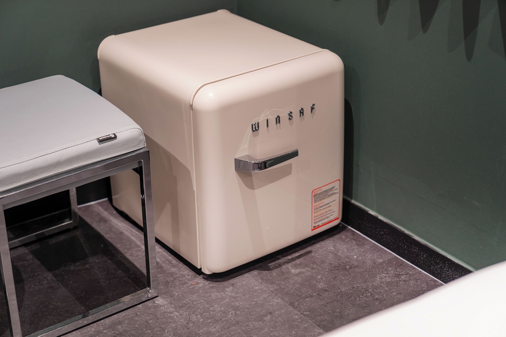 Un refrigerador de estilo vintage con el nombre de marca 'WINSAF' en el frente, ubicado junto a una mesa en una habitación con piso de baldosas y paredes verdes.