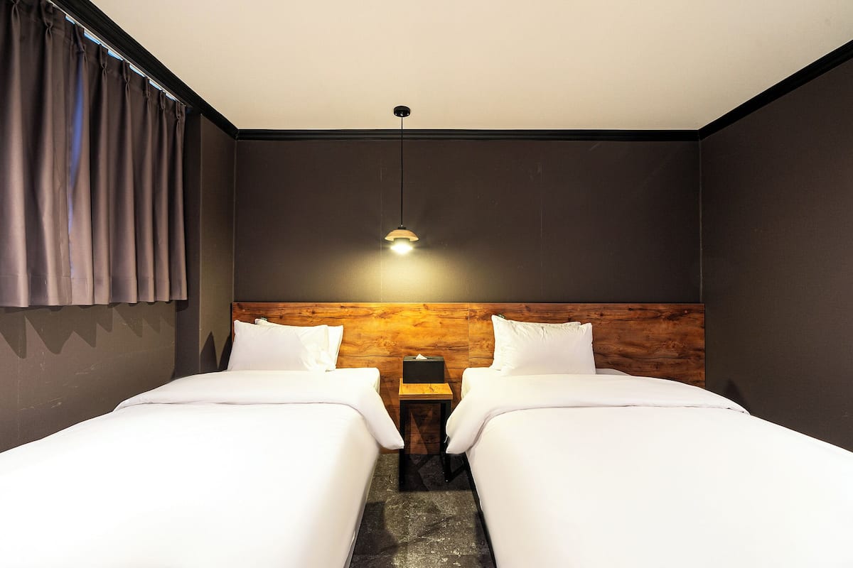 Een hotelkamer met twee enkele bedden, een houten hoofdbord, een nachtkastje met een lamp en een raam met gordijnen.