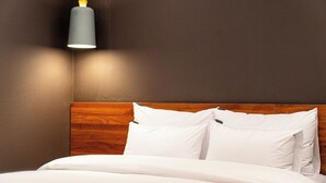 Una cama bien hecha con ropa blanca y almohadas, cabecera de madera y una lámpara moderna fijada a la pared.